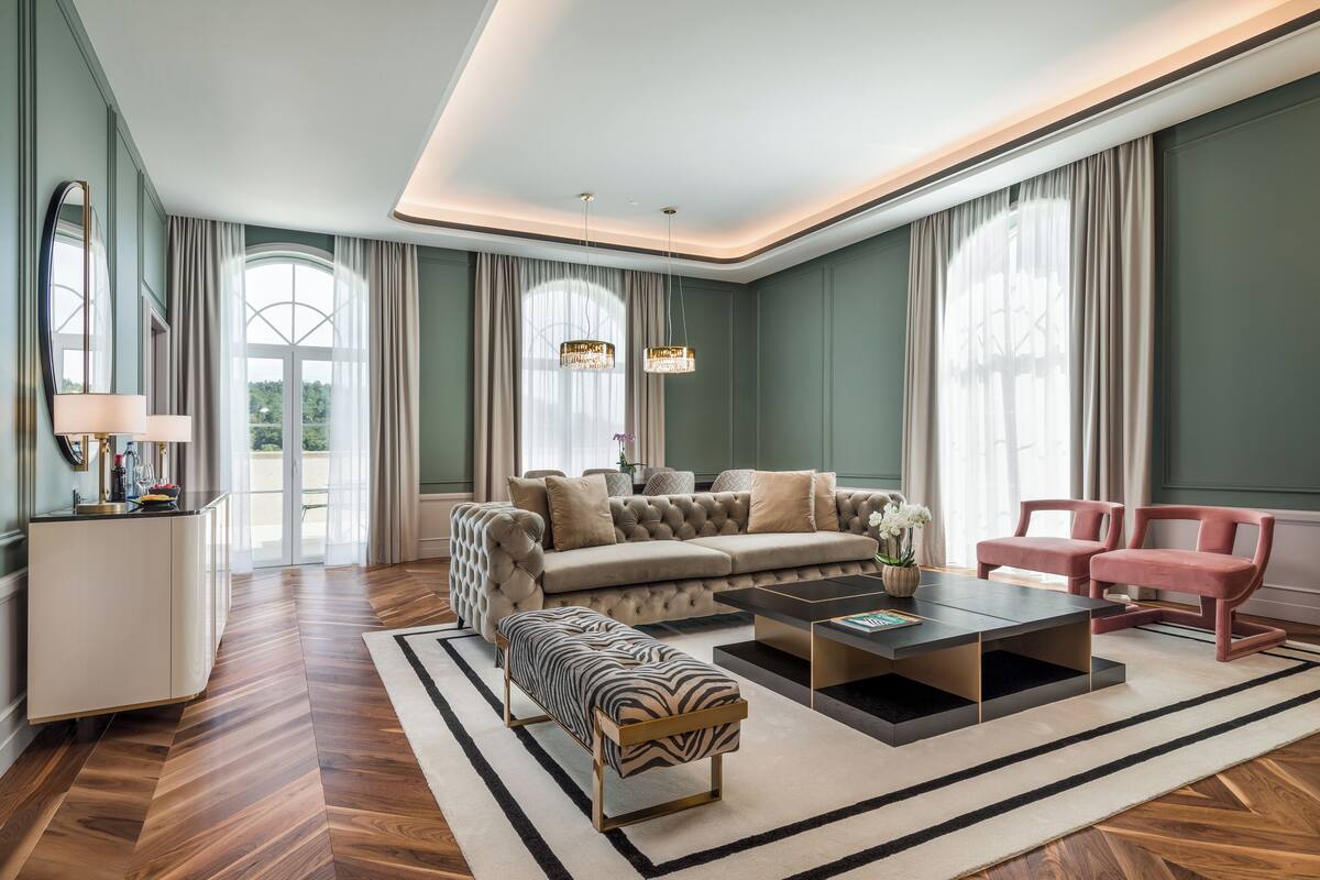 Een ruime woonkamer met een grote bank, een salontafel en een lange bank. Er is een raam met gordijnen, een kroonluchter en een spiegel aan de muur.