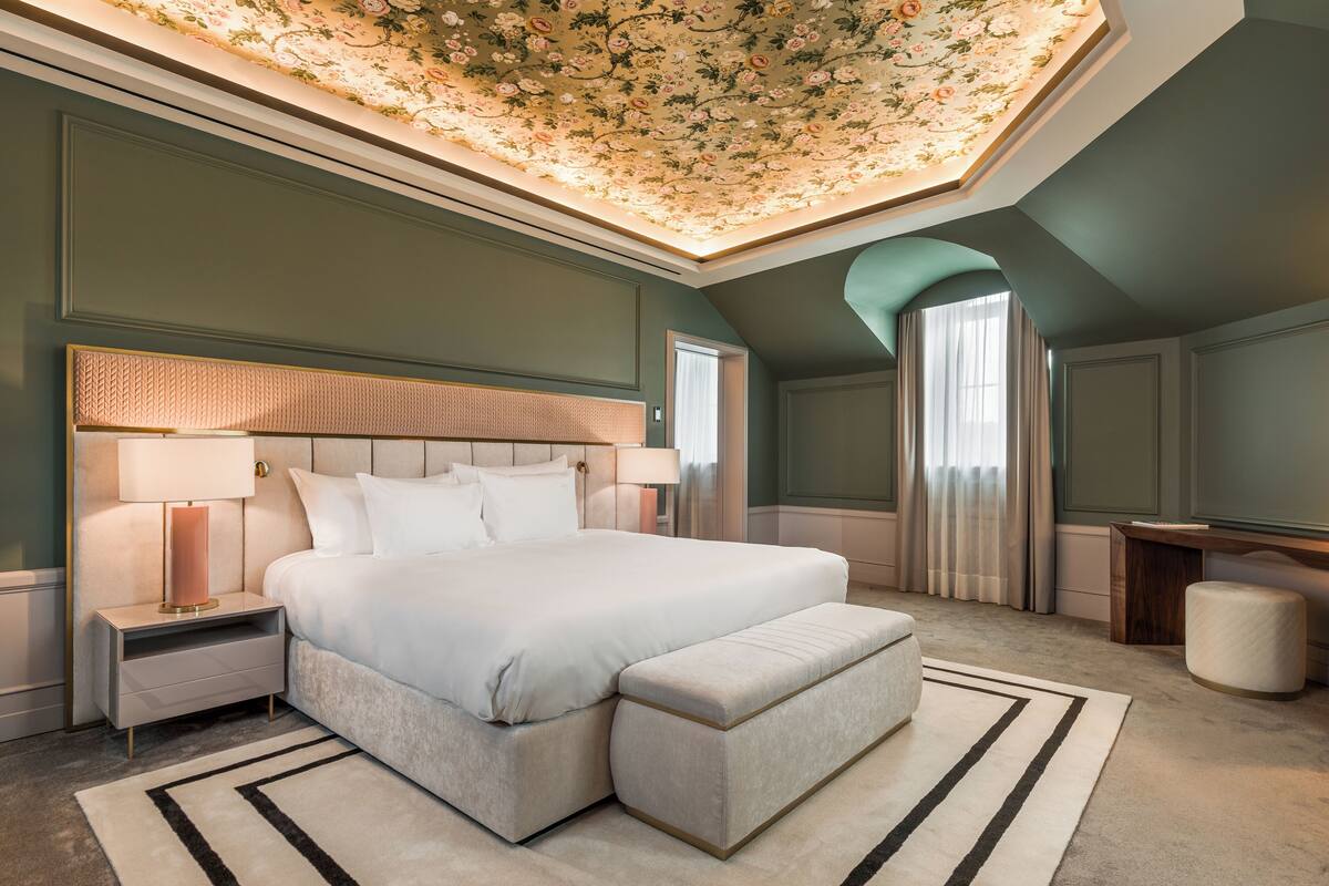 Een slaapkamer met een groot bed, nachtkastjes, een bankje, een bureau en een plafond met bloemmotieven.