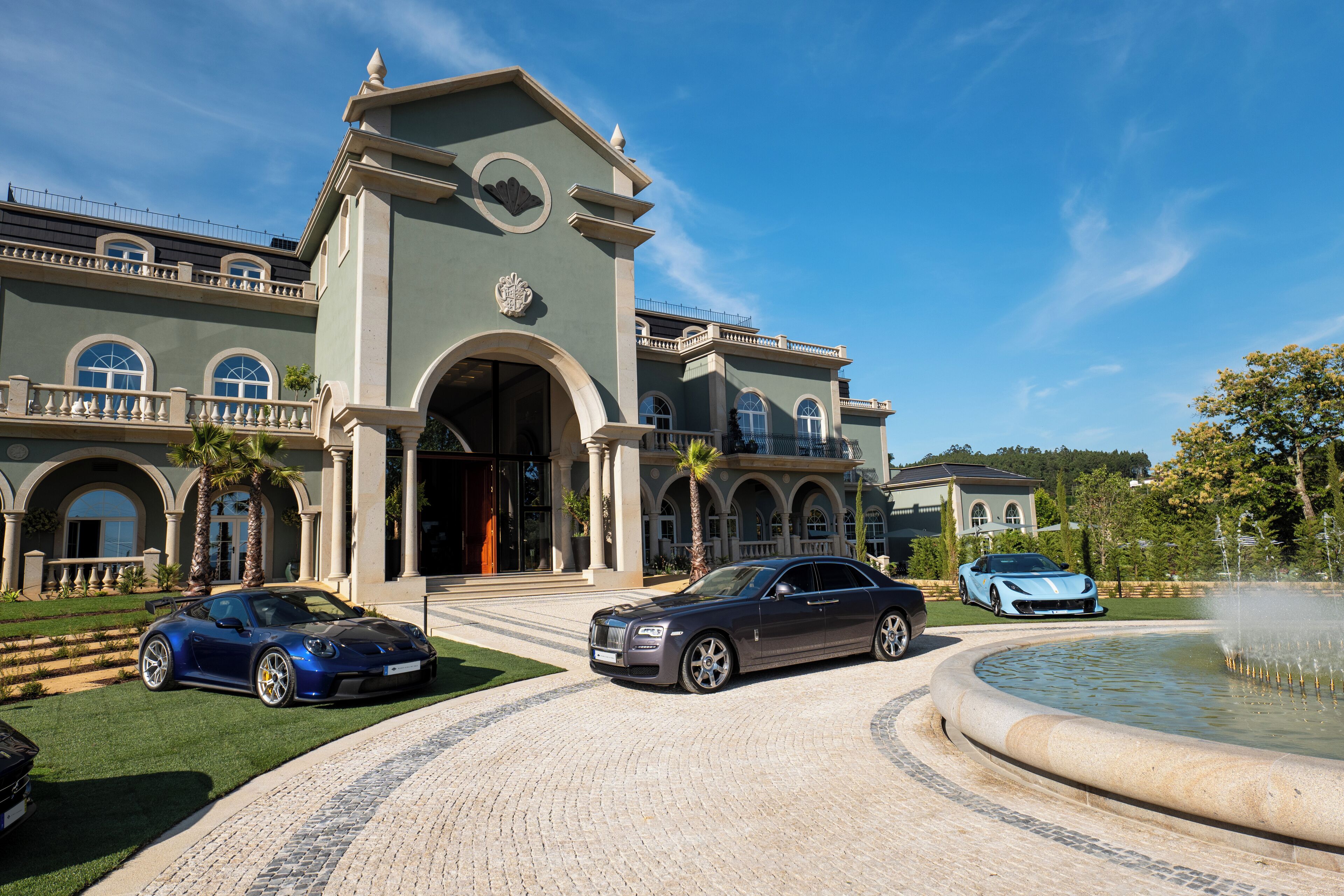 A grand building with a fountain, a driveway, and two luxury cars parked in front.