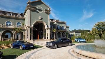 A grand building with a fountain, a driveway, and two luxury cars parked in front.