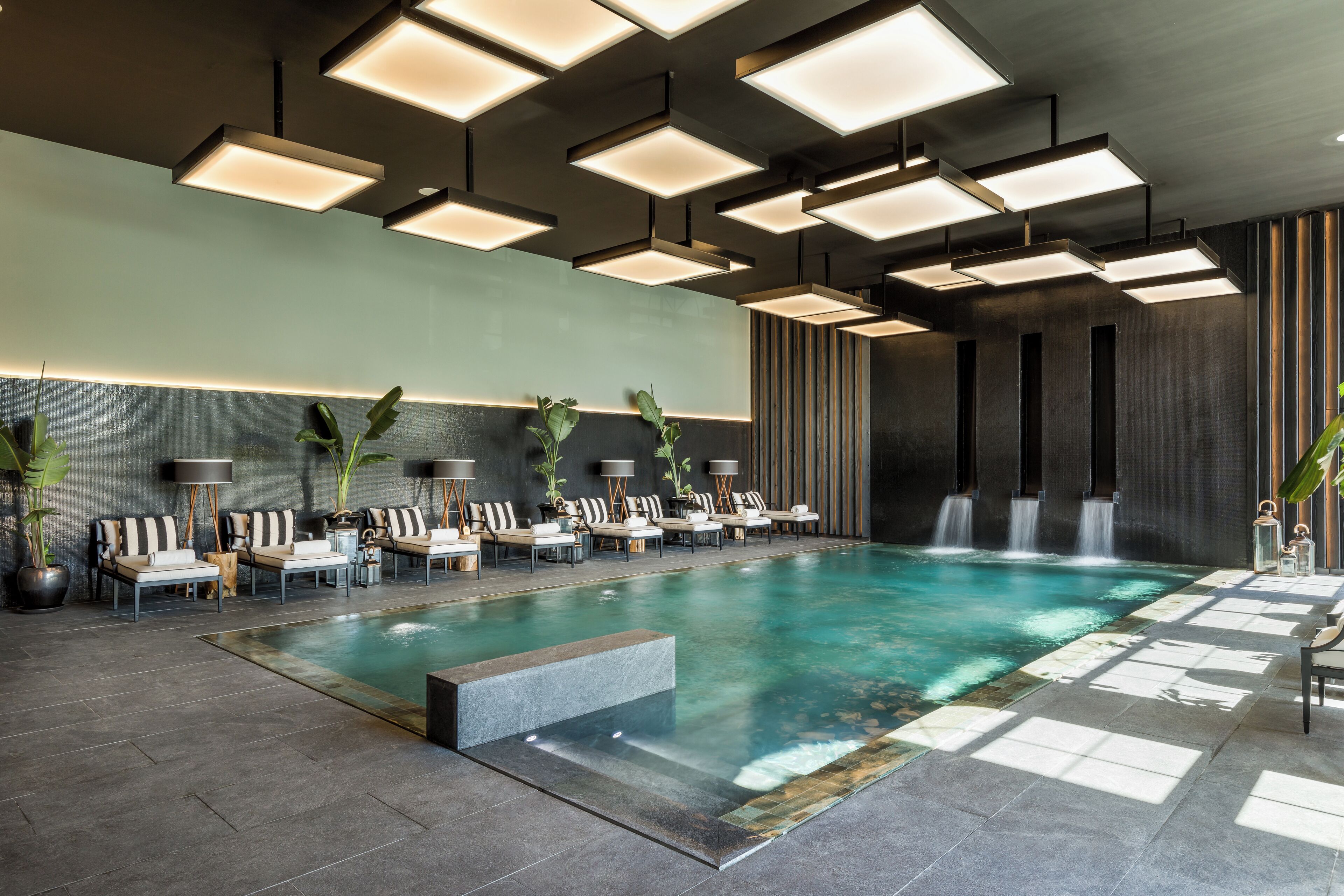 A modern indoor pool area with multiple lounge chairs and tables, a central pool, and a waterfall feature.