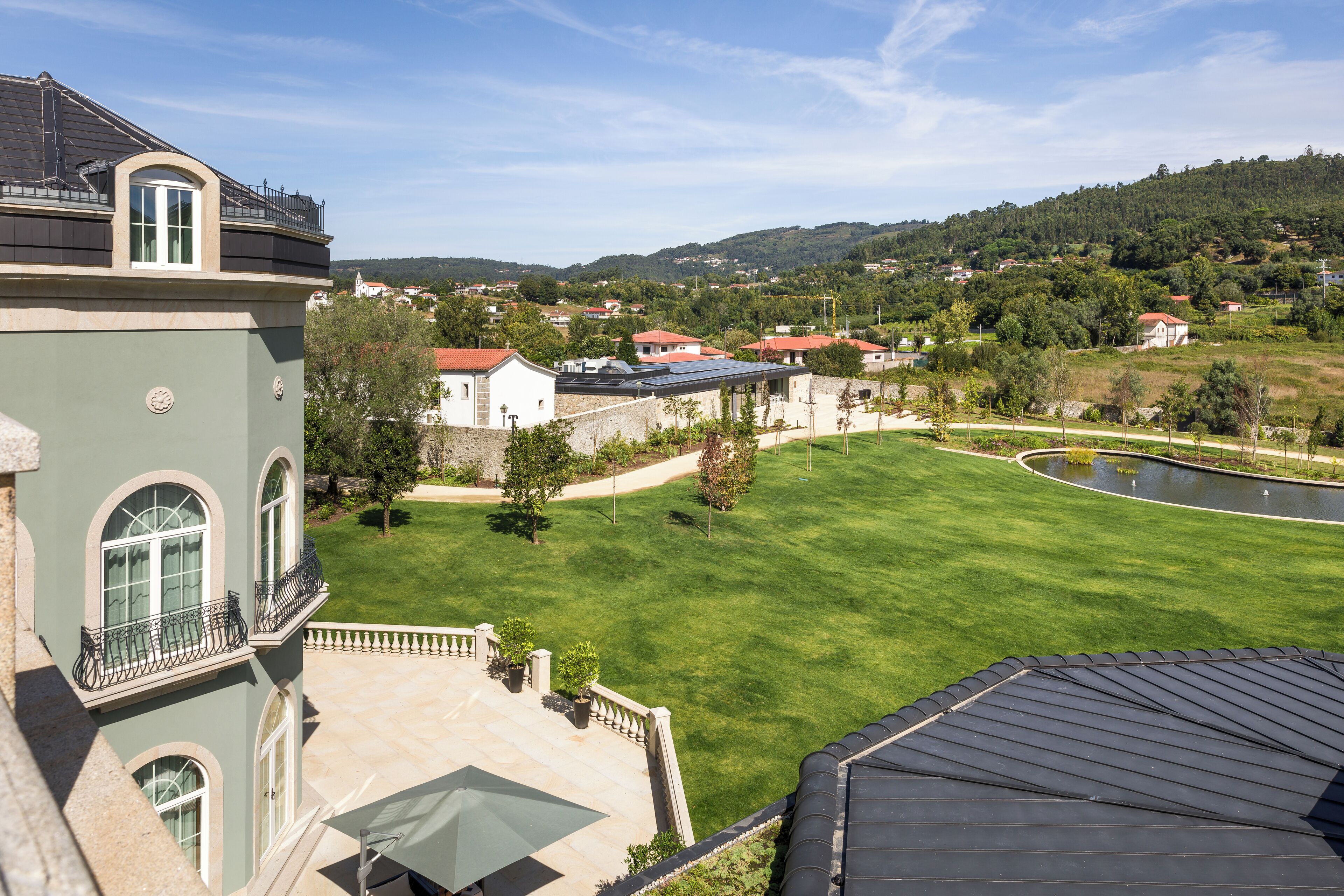 A large building with a green facade, a courtyard with a lawn, and a view of a hilly landscape in the background.