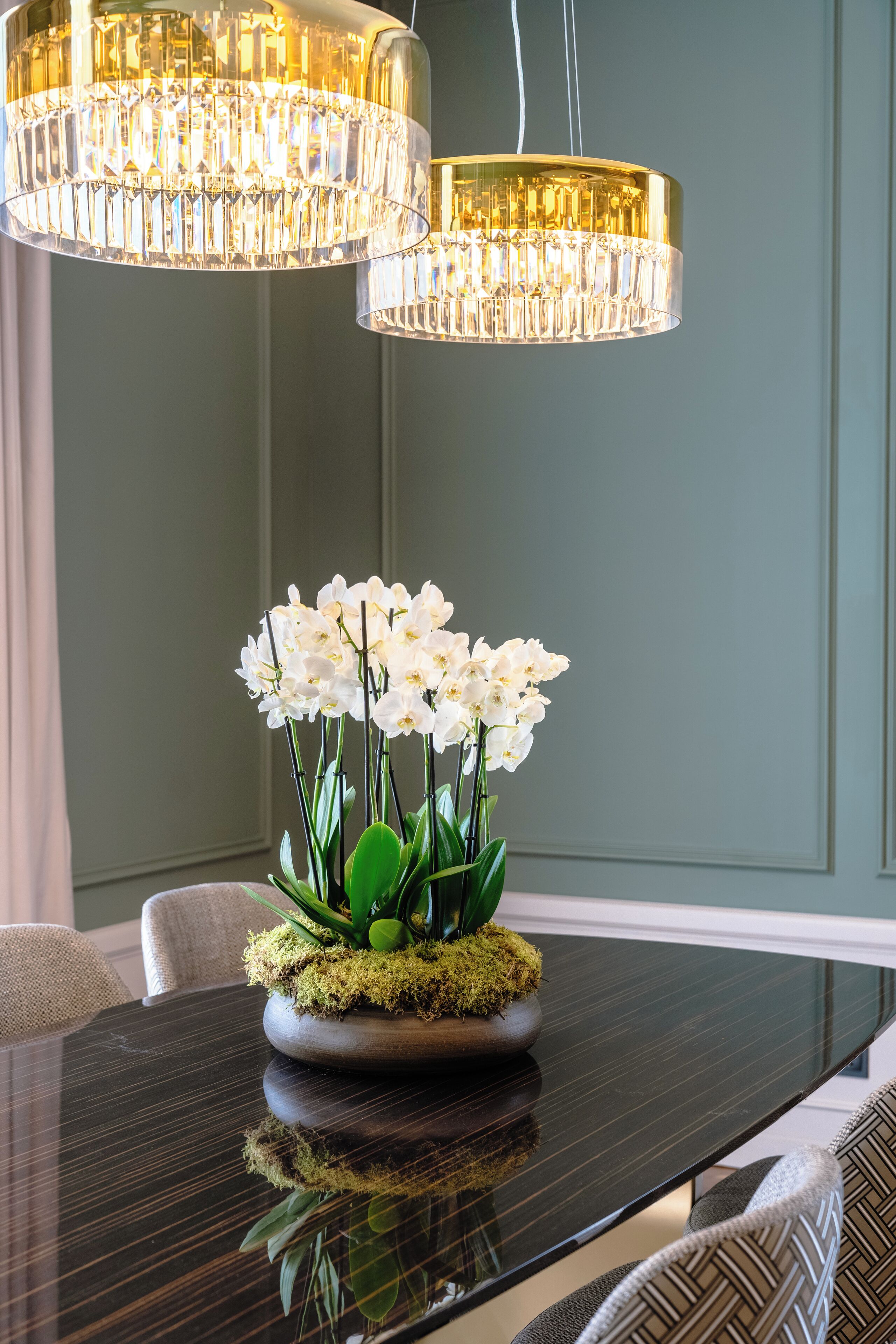 A dining table with a reflective surface, a floral centerpiece, and two hanging chandeliers.