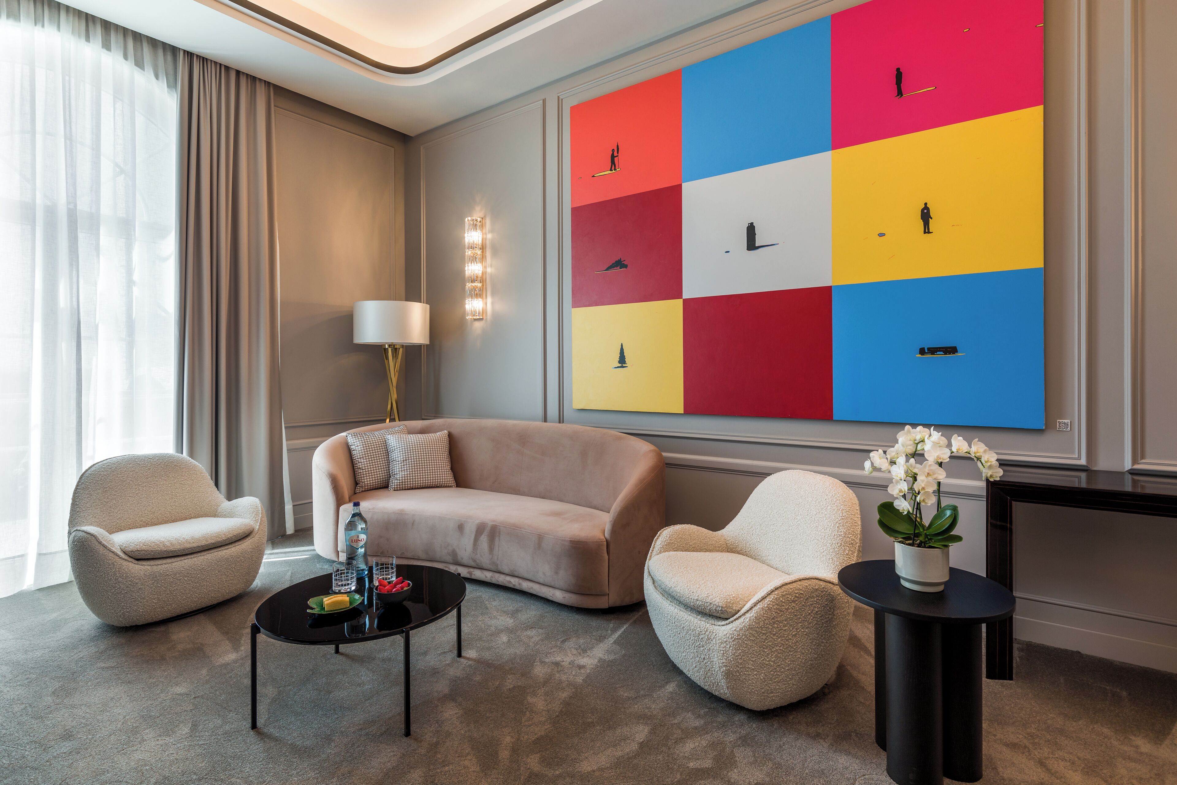 Uma sala de estar moderna com um sofá bege, duas poltronas, uma mesa de centro e um quadro abstrato colorido na parede.