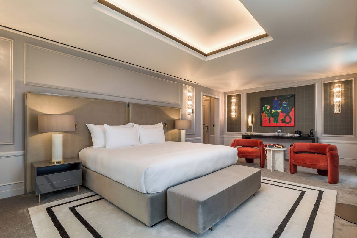 Een moderne hotelkamer met een groot bed, bedlampen, een kleine tafel, twee stoelen en een schilderij aan de muur.
