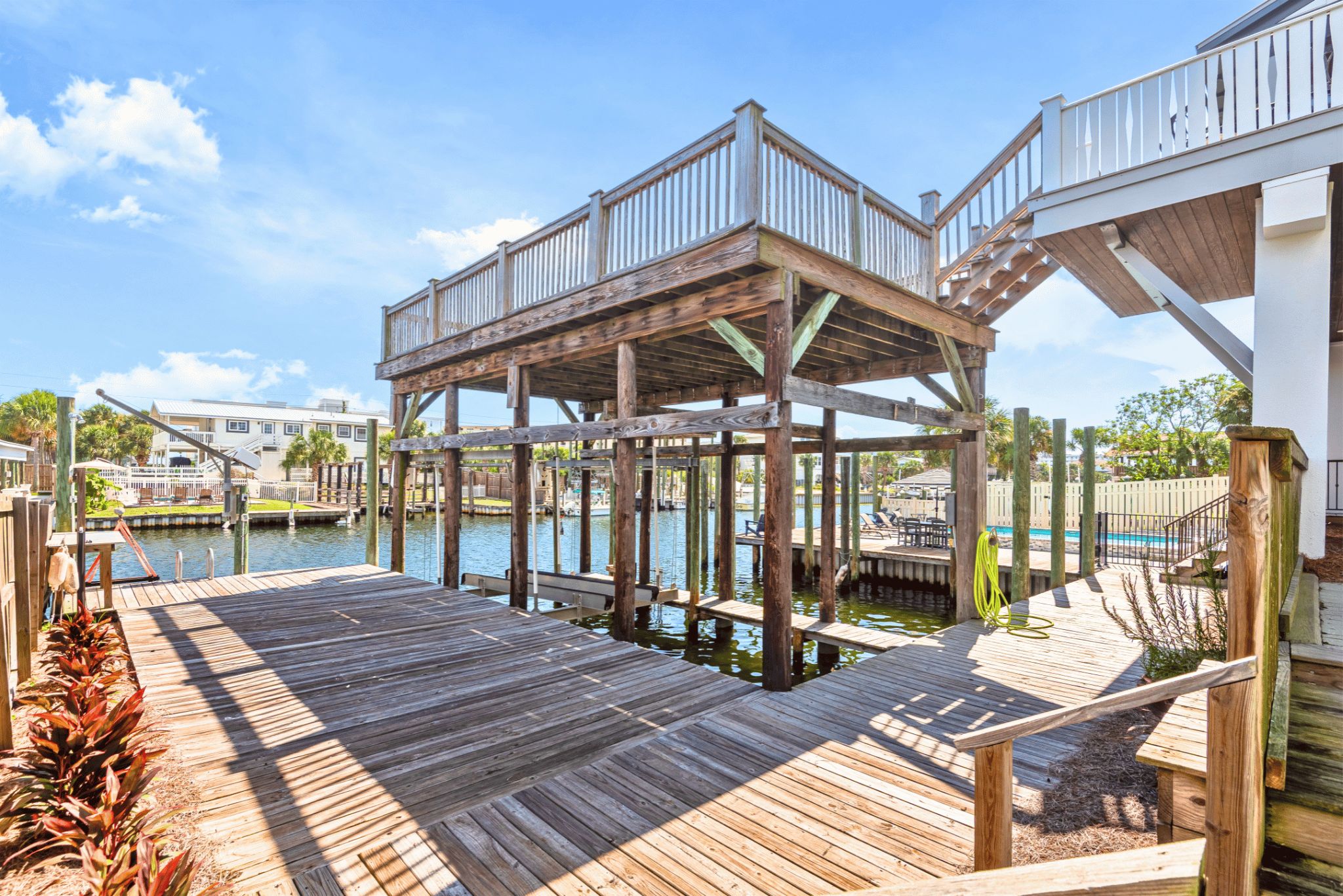 A wooden dock extending over water with a covered seating area and a pool in the background.
