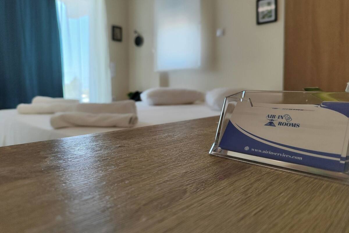 A hotel room with a neatly made bed, a wooden table with a clear plastic sign, and a window with curtains.