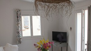 A living room with a sofa, a coffee table with flowers, a television on a stand, and a large window with curtains. - Maison de La Porte des Loups au Pied du Ventoux à Ste Colombe (Bédoin)
