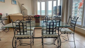 Een eetgedeelte met een glazen tafel, metalen stoelen, een bank, een televisie en uitzicht op zee.