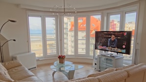 Een woonkamer met een bank, een glazen salontafel, een televisie en uitzicht op gebouwen door grote ramen.