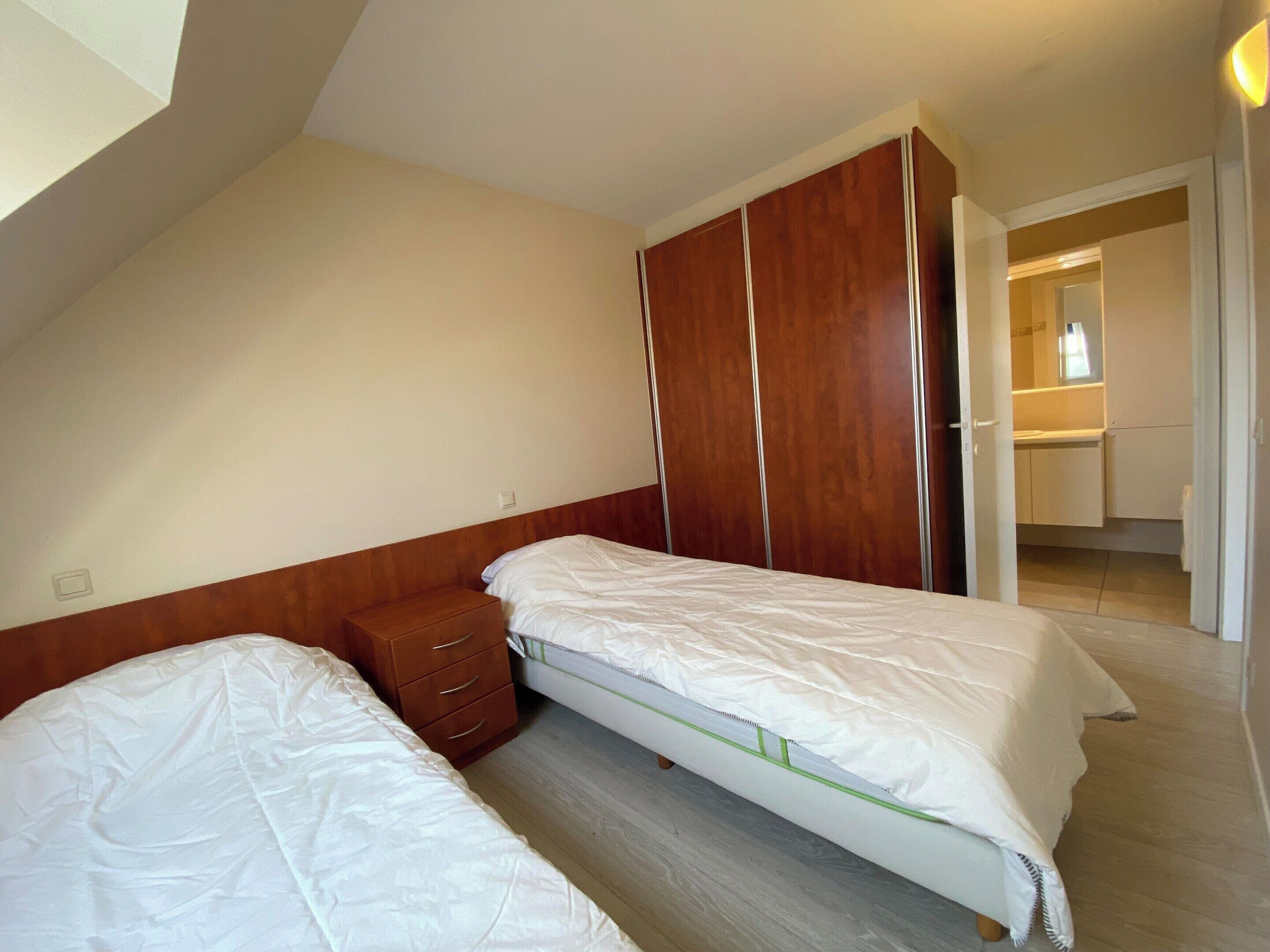 A bedroom with two single beds, a wooden wardrobe, and a bathroom visible through an open door.