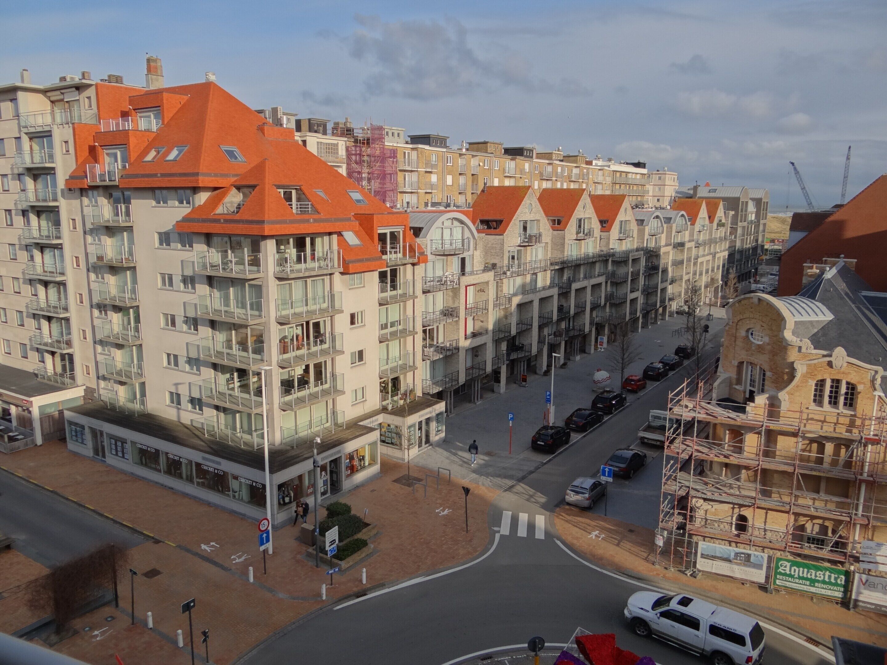 A multi-story building with a red roof, balconies, and a street view with cars and a pedestrian crossing.