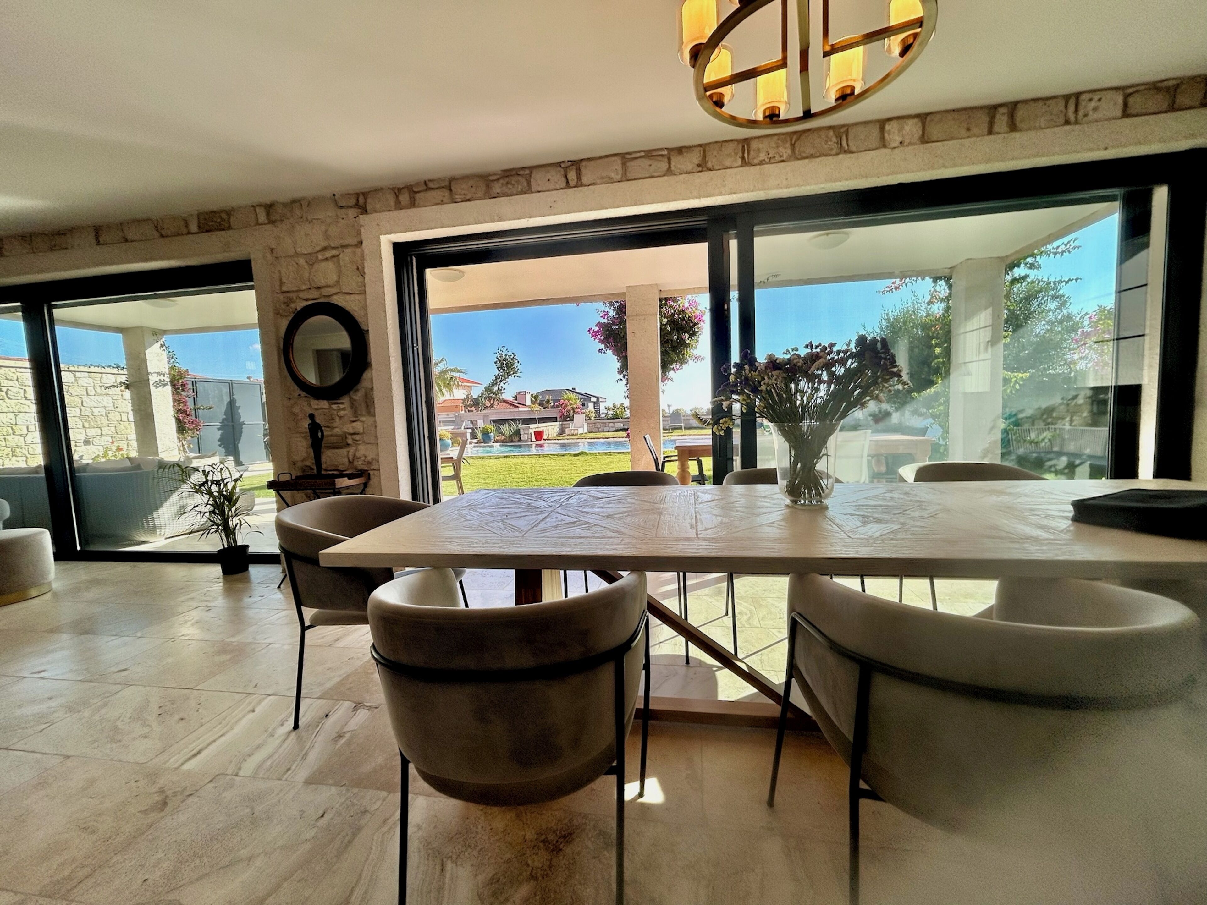 Ein moderner Essbereich mit einem großen Tisch und Stühlen, einer Blumenvase und Blick auf Pool und Garten durch große Glasüren.