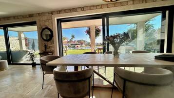 Ein moderner Essbereich mit einem großen Tisch und Stühlen, einer Blumenvase und Blick auf Pool und Garten durch große Glasüren.
