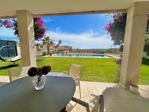 Une terrasse avec une table, des chaises et un vase de fleurs. On aperçoit une piscine au fond.