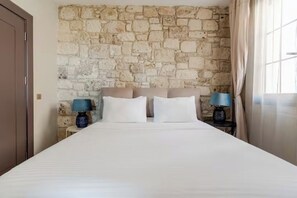 A bedroom with a stone wall, a bed with white bedding, two bedside tables with lamps, and a window with curtains.