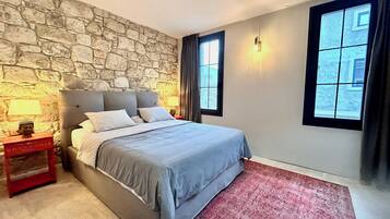 A bedroom with a stone wall, a bed with grey bedding, a red nightstand with a lamp, and a window with curtains.