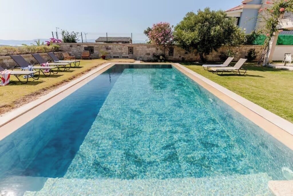 A rectangular swimming pool with clear water, surrounded by a stone wall and a grassy area with lounge chairs.