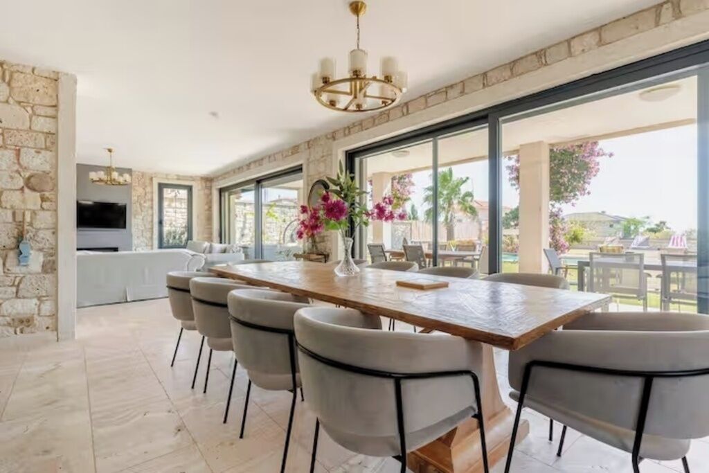 A spacious dining area with a long table and chairs, a chandelier, and a view of the outdoors.