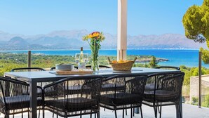 A table set for two with a bottle of Gerald Stay wine, a vase with flowers, and a basket of fruit. The table is outdoors with a view of a body of water and mountains in the background. - Villa Panoramica - modern villa in Alcudia (Alcudia)