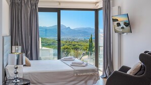 A bedroom with a large bed, a chair, a bedside table, and a view of a mountainous landscape. - Villa Panoramica - modern villa in Alcudia (Alcudia)
