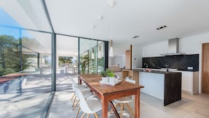 A modern kitchen with a large island, a dining area with a wooden table and white chairs, and large glass windows offering a view of the outdoors. - Villa Sky - private swimming pool and terrace (Alcudia)