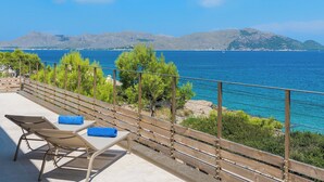 Ein Balkon mit zwei Liegestühlen und einem Holgitter, der Blick auf eine Küstenlandschaft mit klarem, blauem Meer und entfernten Bergen freigibt.