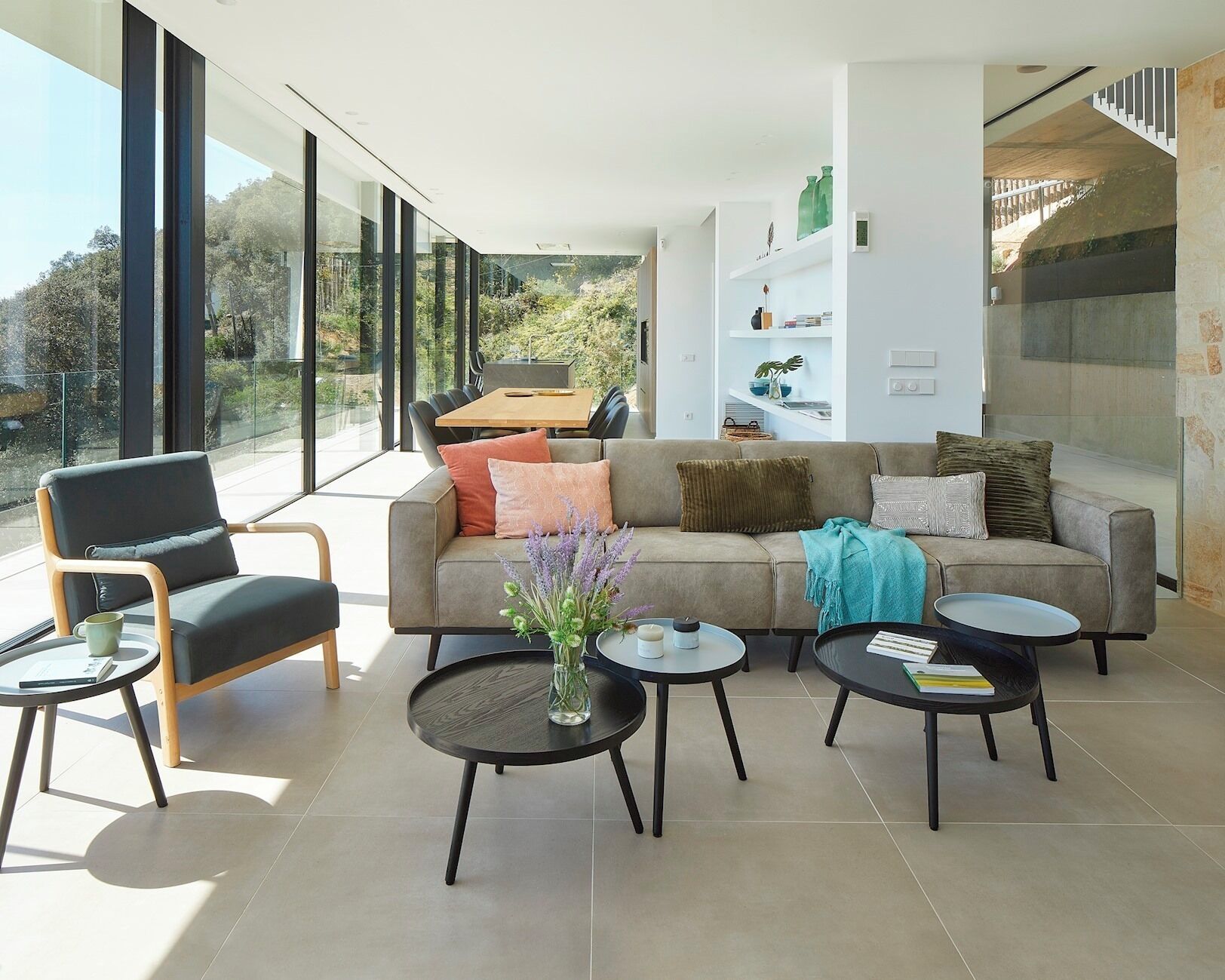 A modern living room with a sofa, two armchairs, and a coffee table. There is a bookshelf with decorative items and a view of the outdoors through large windows.