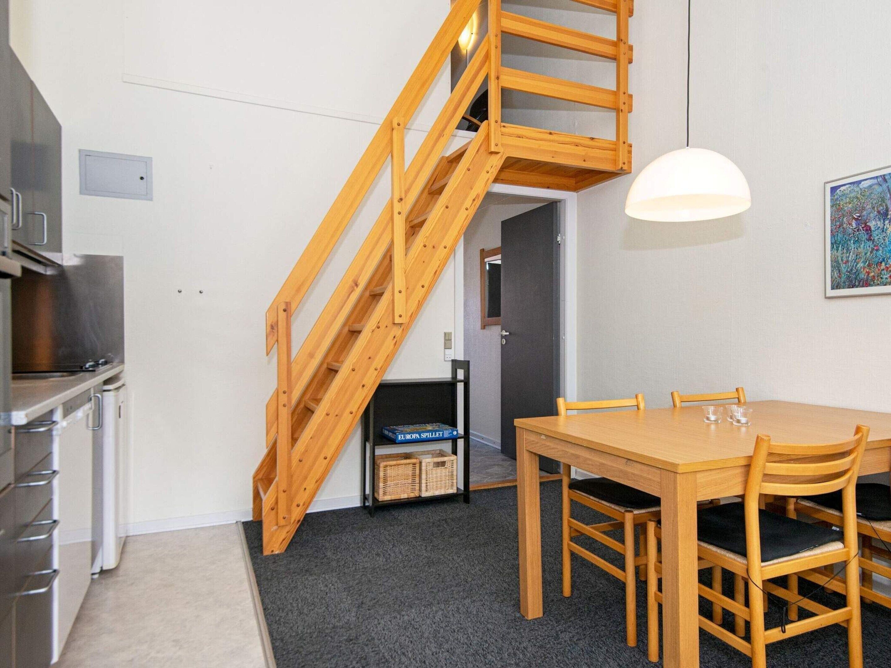 A compact kitchen with a wooden staircase leading to a door, a dining table with chairs, and a hanging light fixture.