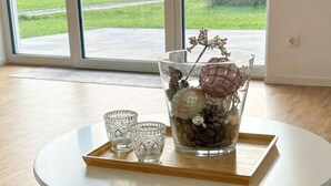 Une table ronde blanche, dotée d’un pied en fil métallique aux motifs géométriques, un vase en verre contenant des fleurs séchées et un plateau sur lequel reposent deux verres, le tout posé sur un tapis gris à texture marquée.