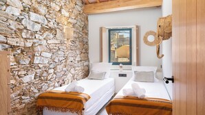 Une chambre avec deux lits, des murs en pierre, un plafond en bois et une fenêtre donnant sur des immeubles.