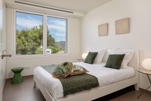 Ein Schlafzimmer mit einem Bett, einem grünen Nachttisch, einem Fenster mit Blick auf Bäume und Hügel sowie zwei an der Wand befestigten Kunstwerken.
