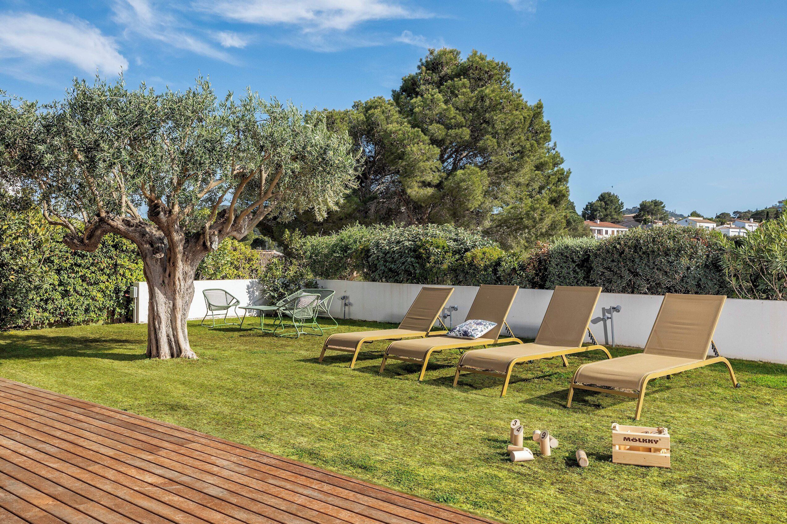 A backyard with a wooden deck, a large tree, and three lounge chairs with cushions.