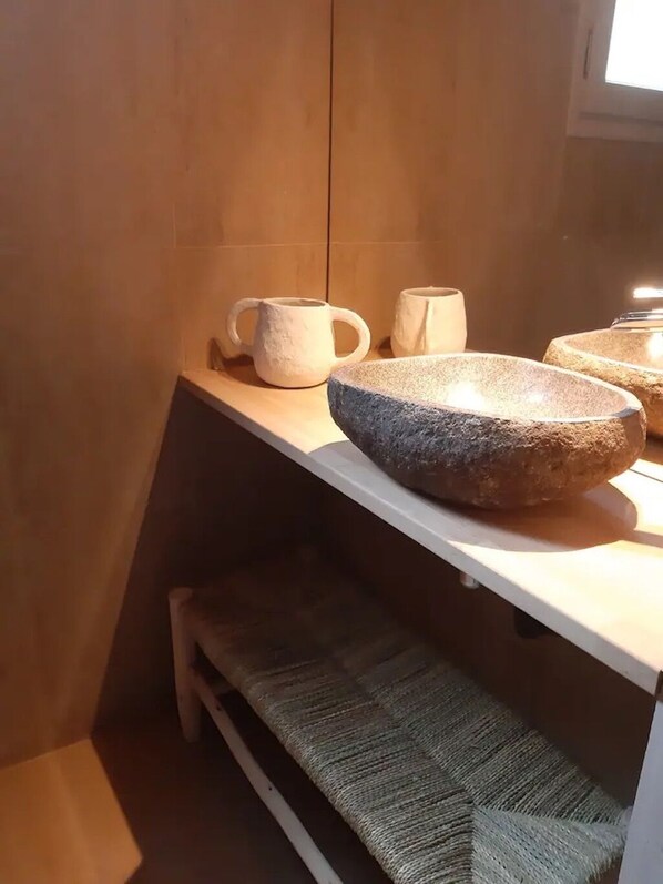 Une salle de bain avec un lavabo en pierre, deux tasses en céramique blanche et un tapis tissé posé sur une étagère.