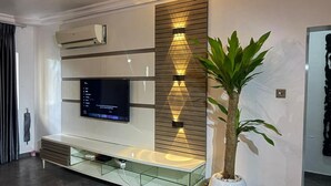A modern living room with a flat-screen TV mounted on a wall, a built-in entertainment unit below, and a decorative wall panel with integrated lighting. - Charming and Spacious 3 Bed in Lekki Phase1 (Lekki)