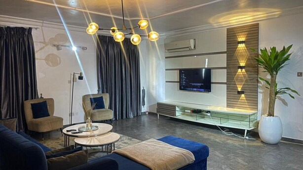 A modern living room with a sofa, armchair, and a coffee table. There is a TV mounted on the wall and a plant in a white pot. - Charming and Spacious 3 Bed in Lekki Phase1 (Lekki)