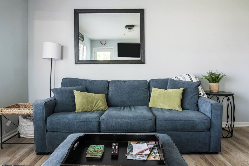 A living room with a grey sofa, a coffee table with books and a remote, a floor lamp, and a small side table with a plant.