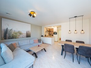 Een moderne woonkamer met een bank, salontafel en een eethoek met stoelen. Achtergrond is een keuken.