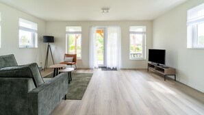 Un salon spacieux avec un canapé gris, une petite table, un téléviseur posé sur un meuble et une porte-fenêtre donnant sur un balcon.