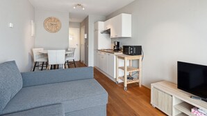 Un salon moderne avec un canapé gris, un coin repas avec une table et des chaises blanches, une cuisine avec des meubles blancs et un micro-ondes noir, et un téléviseur posé sur un meuble en bois.