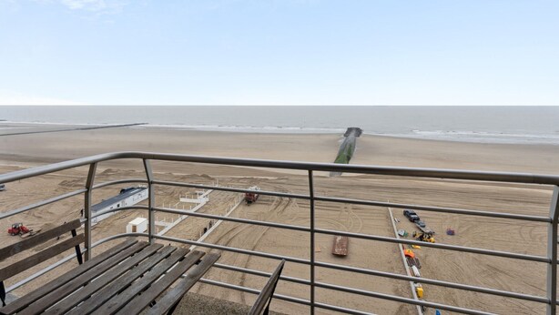 Une vue sur la plage depuis un balcon doté d’une rambarde en métal, avec une plage de sable et une longue jetée qui s’avance dans la mer.