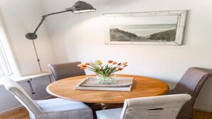 A dining area with a round wooden table, four upholstered chairs, a vase with flowers, a framed picture on the wall, and a floor lamp.