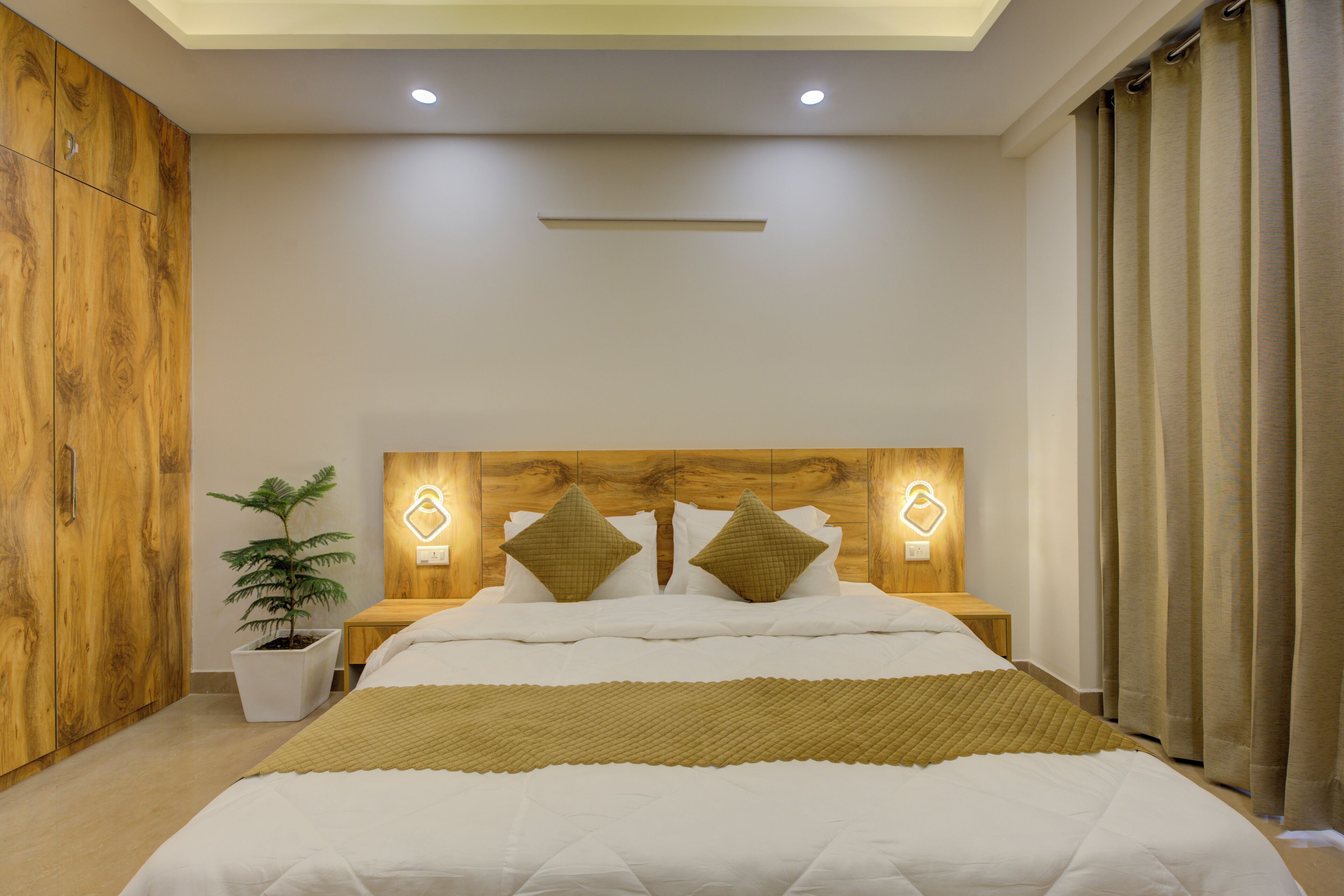 Un dormitorio con cabecera de madera, dos mesitas de noche con lámparas, una planta en maceta y una cama grande con colcha acolchada.