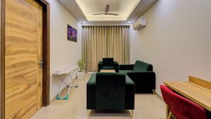 Un espace de vie compact doté d’une porte en bois, d’un ventilateur de plafond, d’une petite table avec une plante, de deux canapés vert foncé, d’un fauteuil rouge et d’une table en bois.