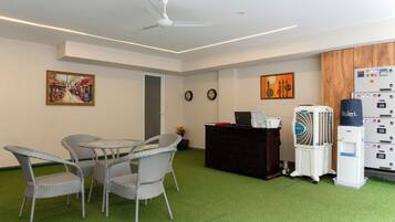 Una habitación con una mesa redonda, sillas de mimbre, un ventilador de techo, un escritorio negro con una computadora y un dispensador de agua, y dos relojes de pared.