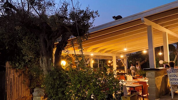 An outdoor patio area with white wicker chairs, a stone planter, and a covered seating area with tables and chairs.