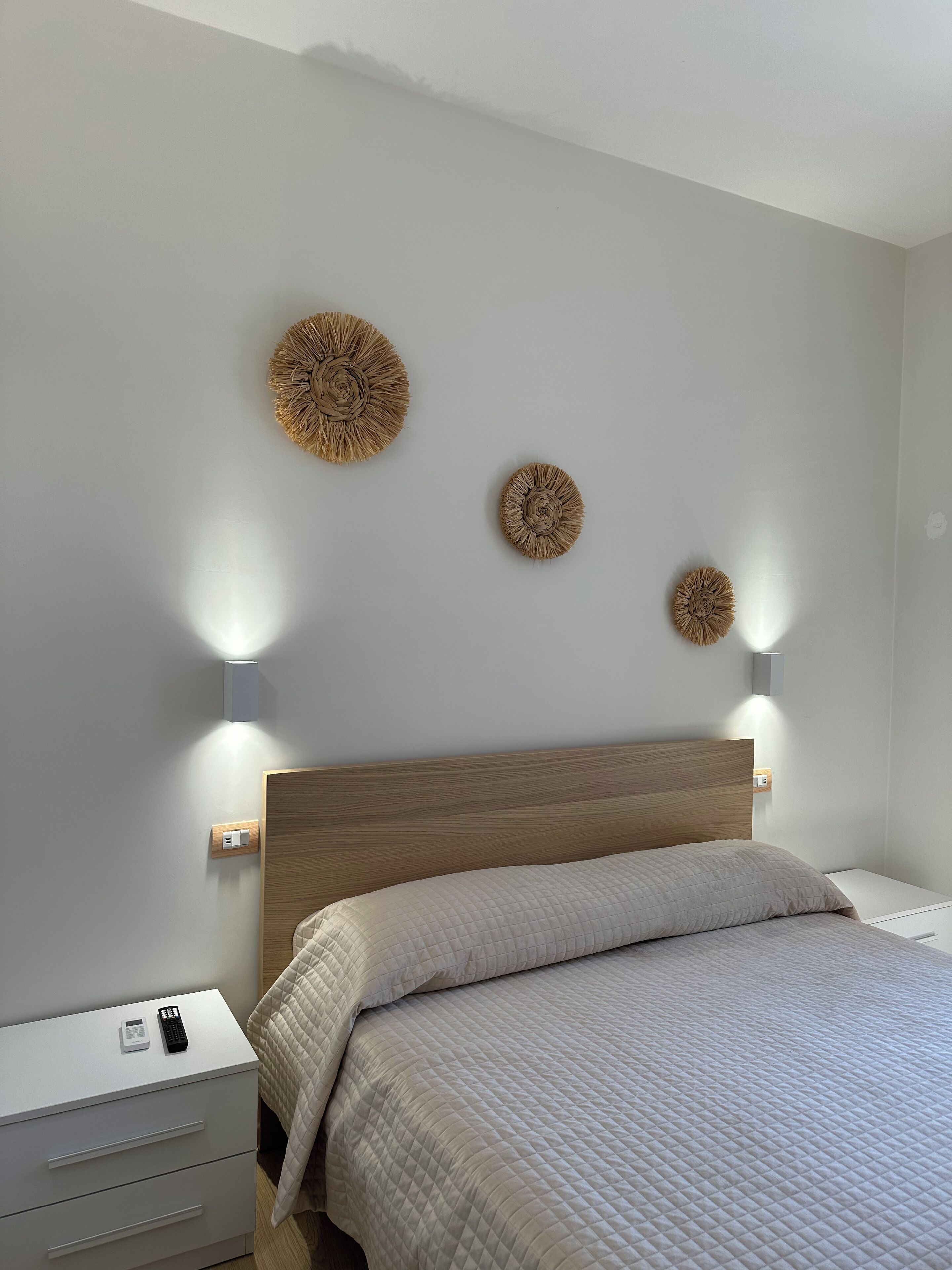 Een slaapkamer met een houten hoofdbord, een bed met een gewatteerde sprei, een nachtkastje met een afstandsbediening en wandlampen.