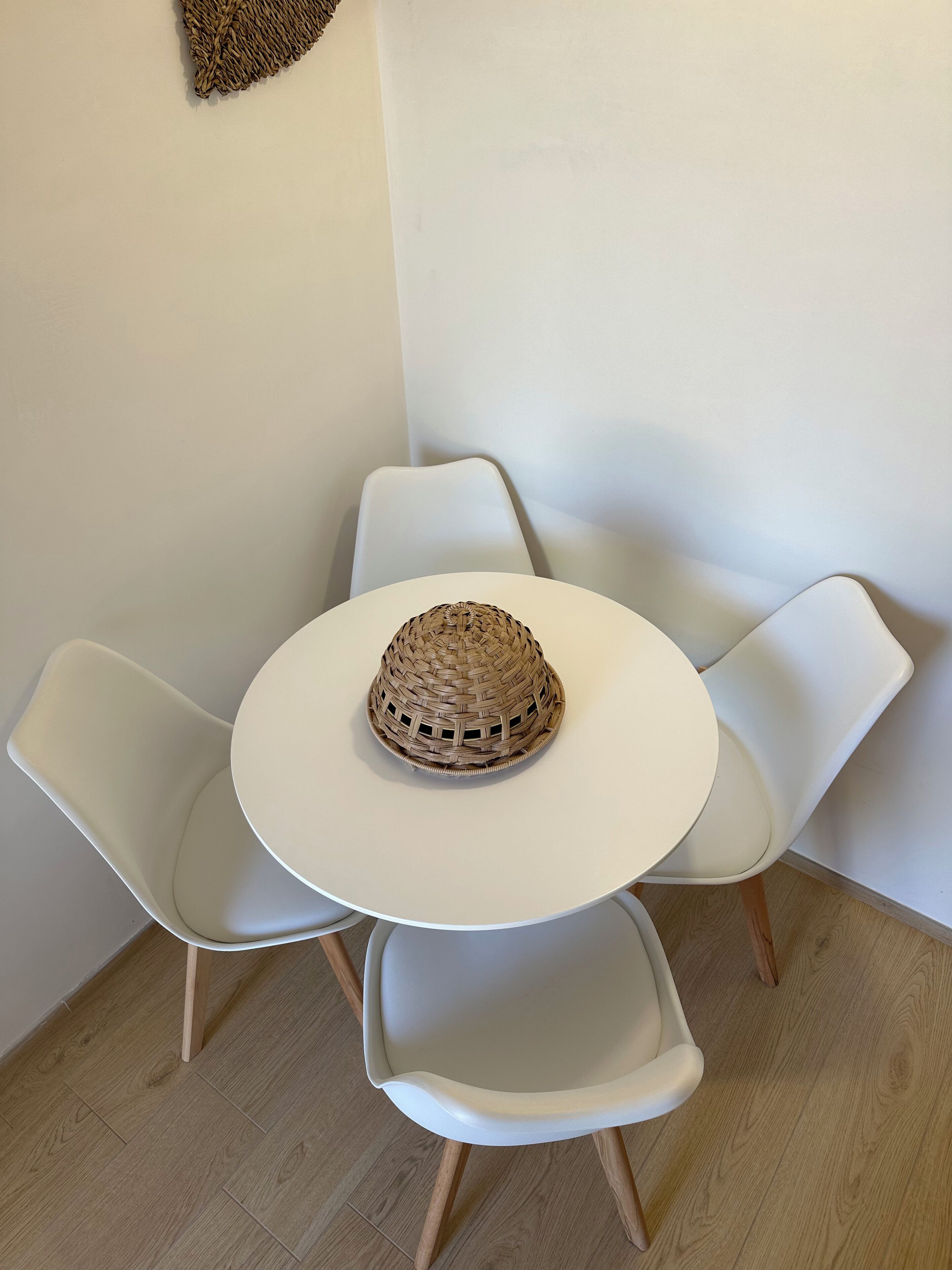 Une table ronde blanche, ornée d’un centre de table en forme de panier tressé, entourée de quatre chaises blanches aux pieds en bois.