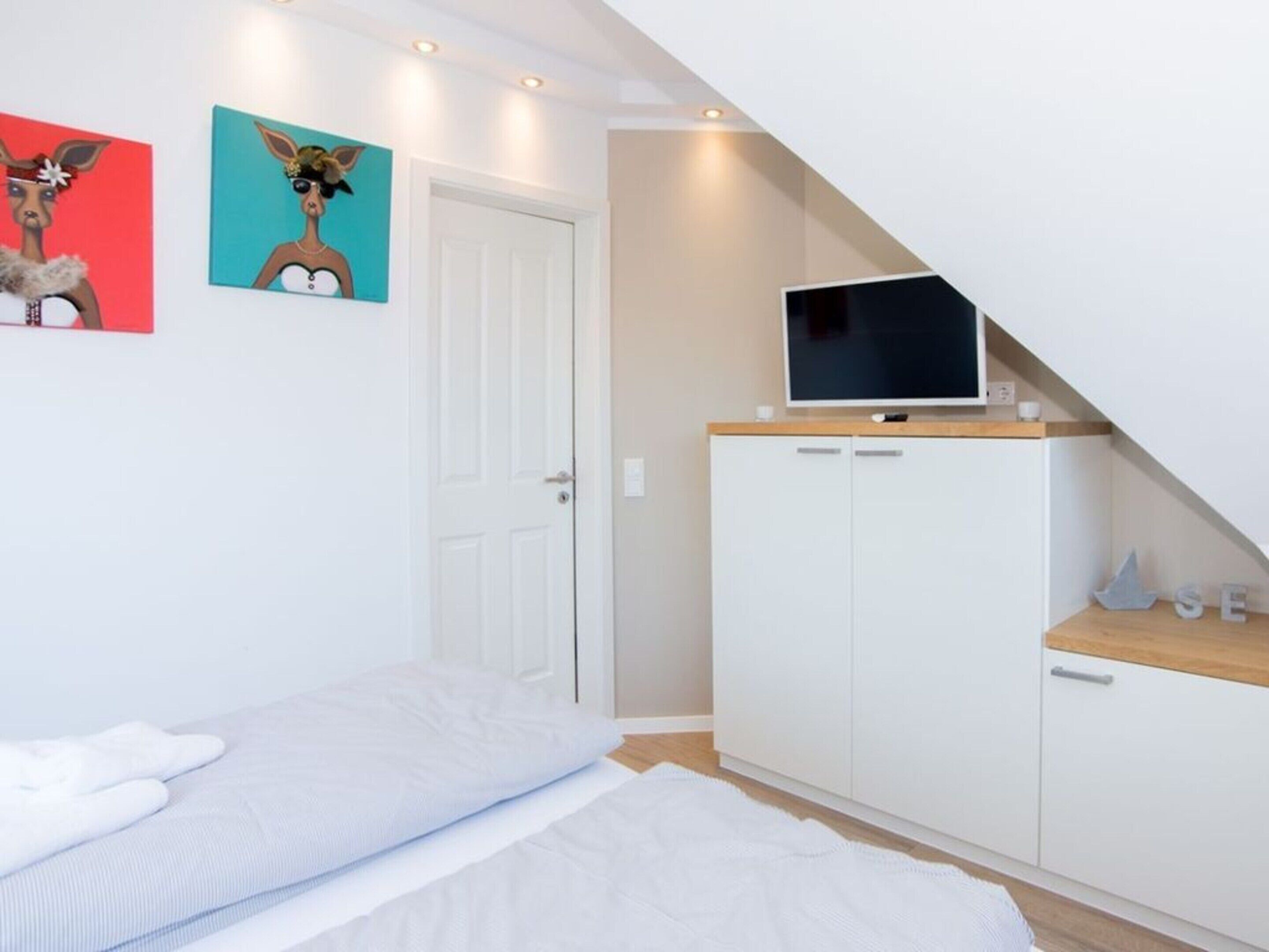 Een slaapkamer met een bed, een witte kledingkast, een televisie erboven en twee kunstwerken aan de muur.