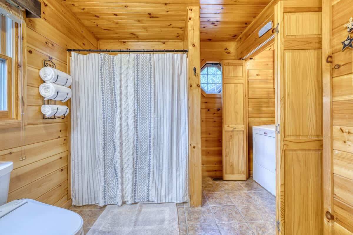 A wooden cabin interior with a bathroom, a shower curtain, a towel rack, a window, and a washing machine.